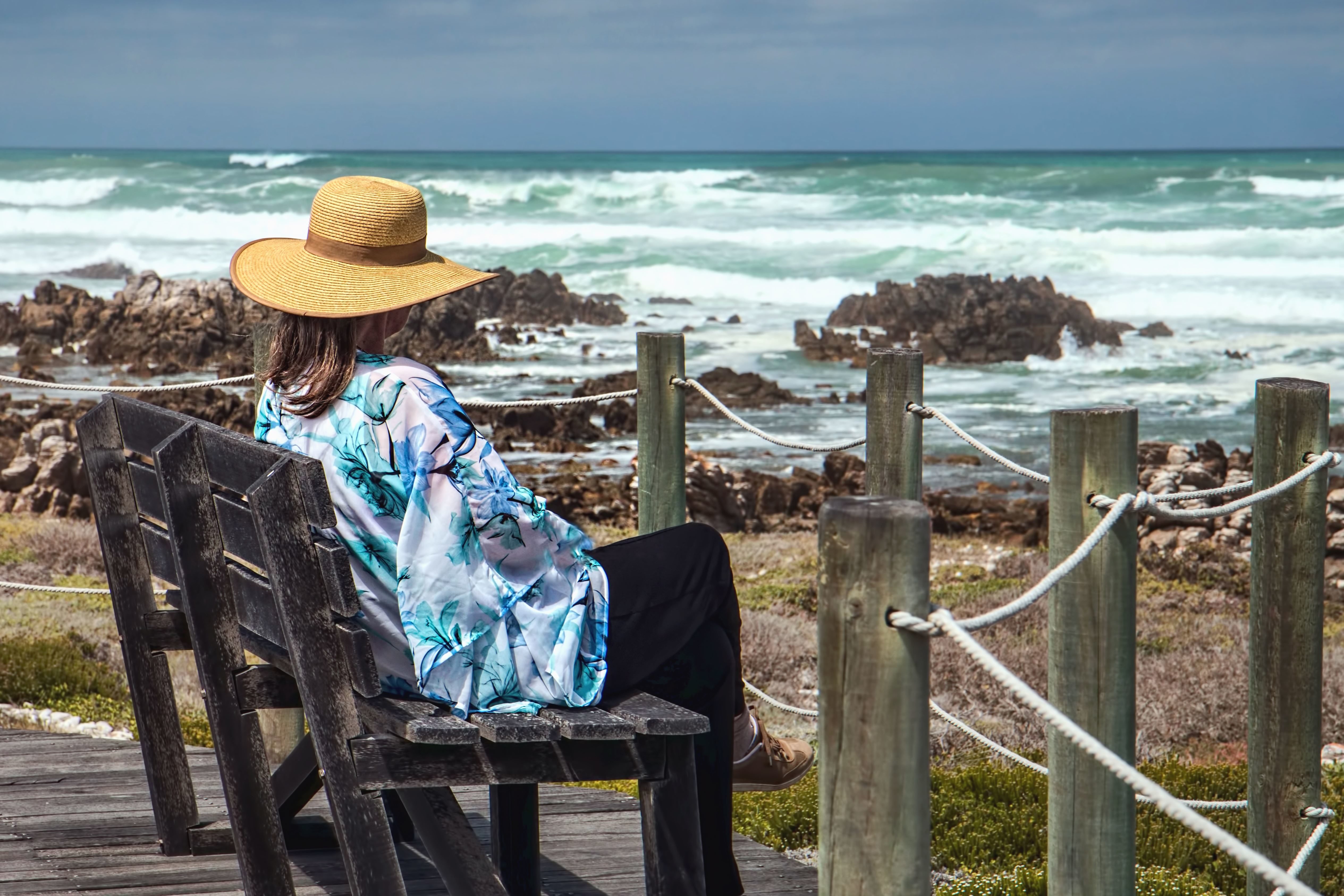 Путешествия размышления. Женщина в шляпе на море. Женщина на скамейке на берегу. Пожилая женщина на берегу. Женщина на берегу океана.