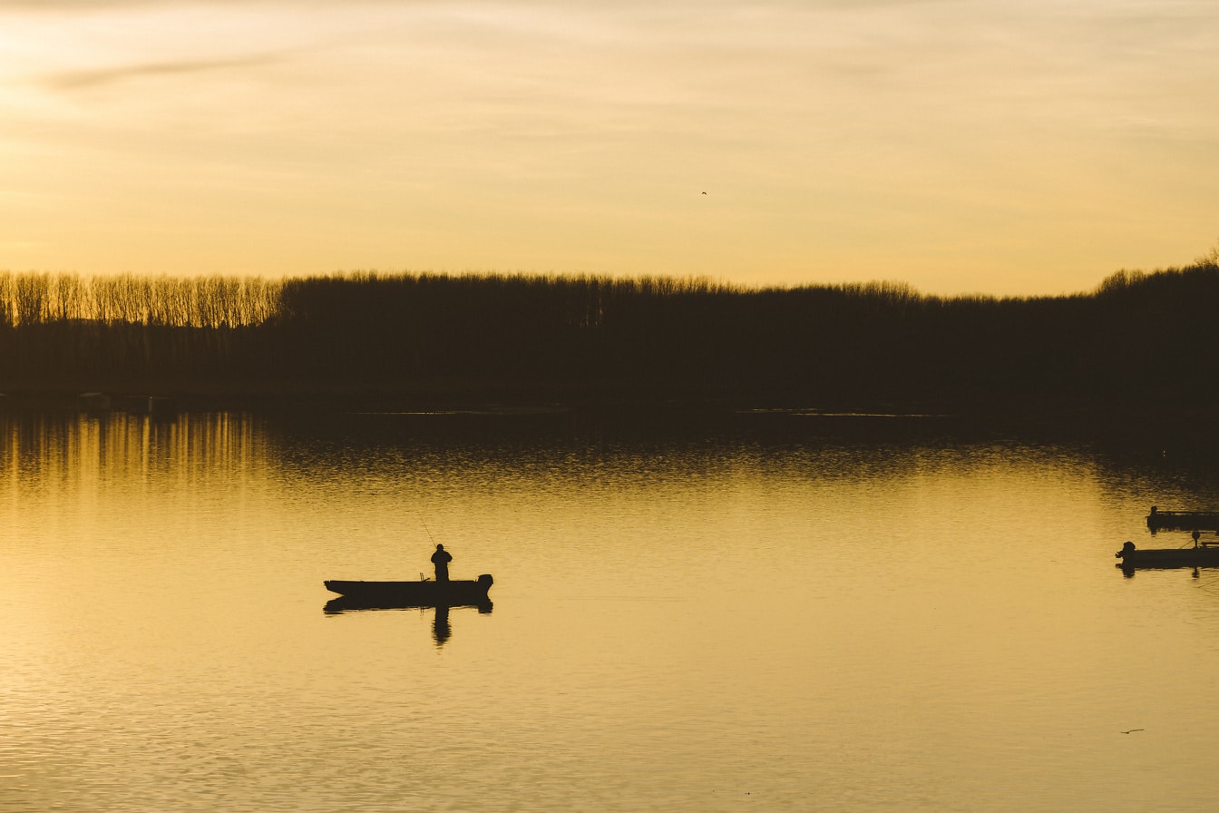 Silueta rybáře v rybářské lodi na klidném jezeře při východu slunce