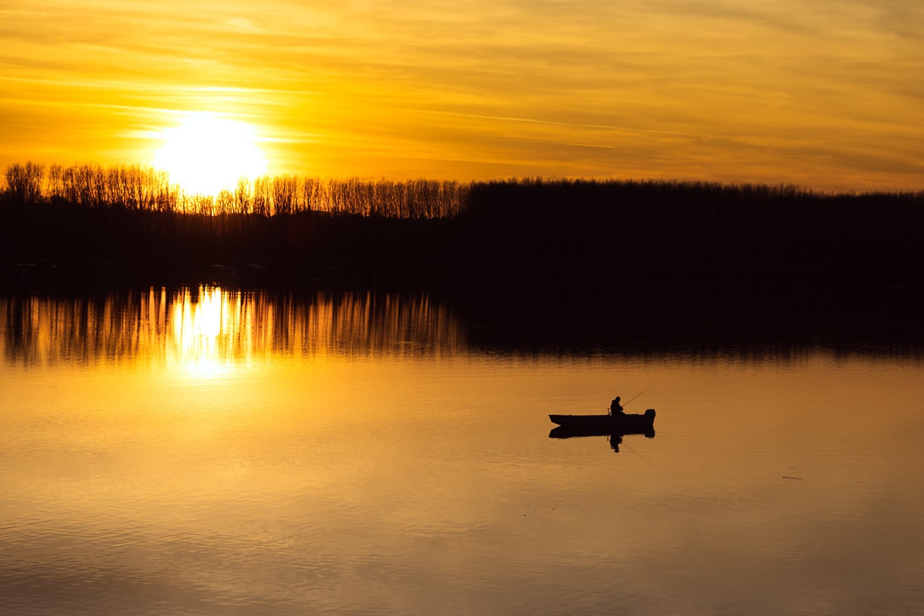 Oranssinkeltainen auringonlasku ja kalastajan siluetti kalastusveneessä