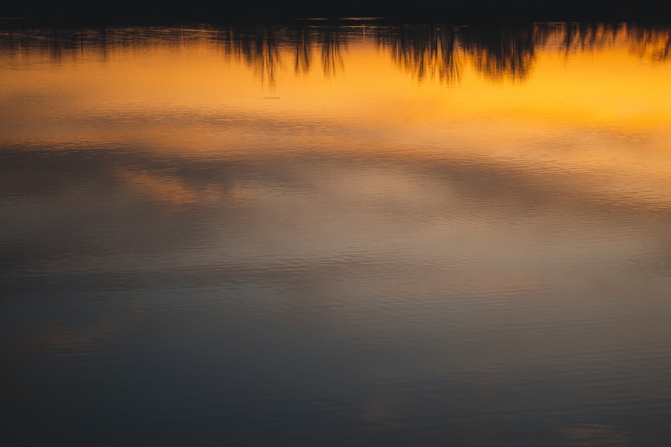 Oranssinkeltainen auringonlaskun taivas heijastuu tyynellä vedellä