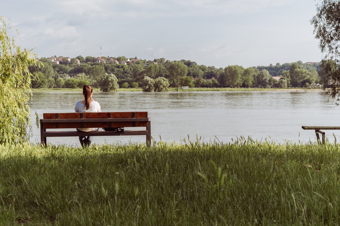 Giovane donna che si siede sulla panchina di legno vicino alla riva del fiume
