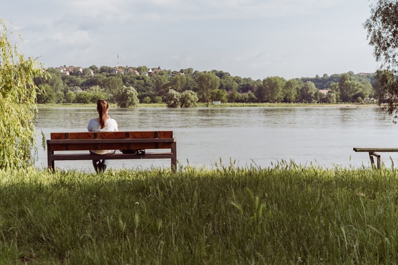 Tânără fată stând pe o bancă de lemn lângă malul râului