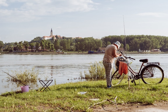 Old woman fishing with fishing rod on riverbank in summer