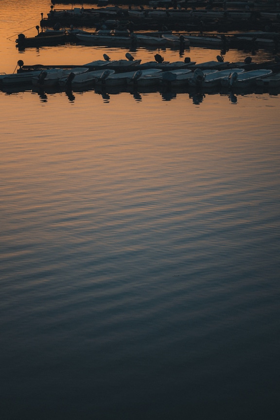 Calm water surface with silhouette of fishing boats in harbor in dusk