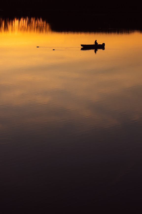 Silhouette of fisherman in fishing boat on calm water with sunset reflections