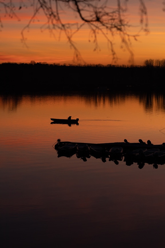 Silhouette of fisherman in fishing boat on calm lake with orange yellow sunset