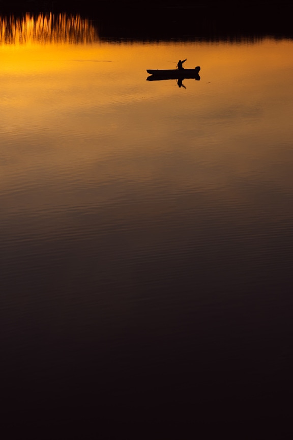 Silhouette of fisherman in fishing boat on calm water at dusk