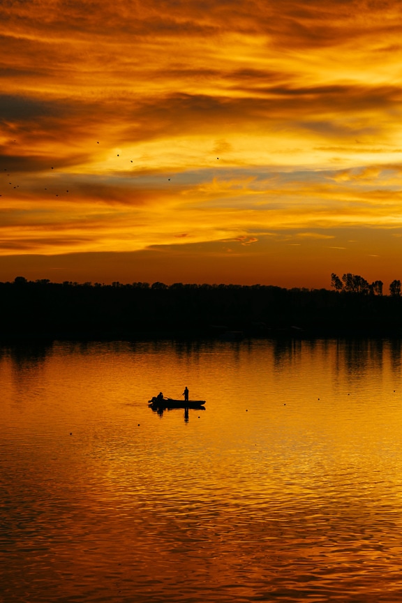Величественный оранжево-желтый закат на берегу озера с силуэтом рыбака в лодке