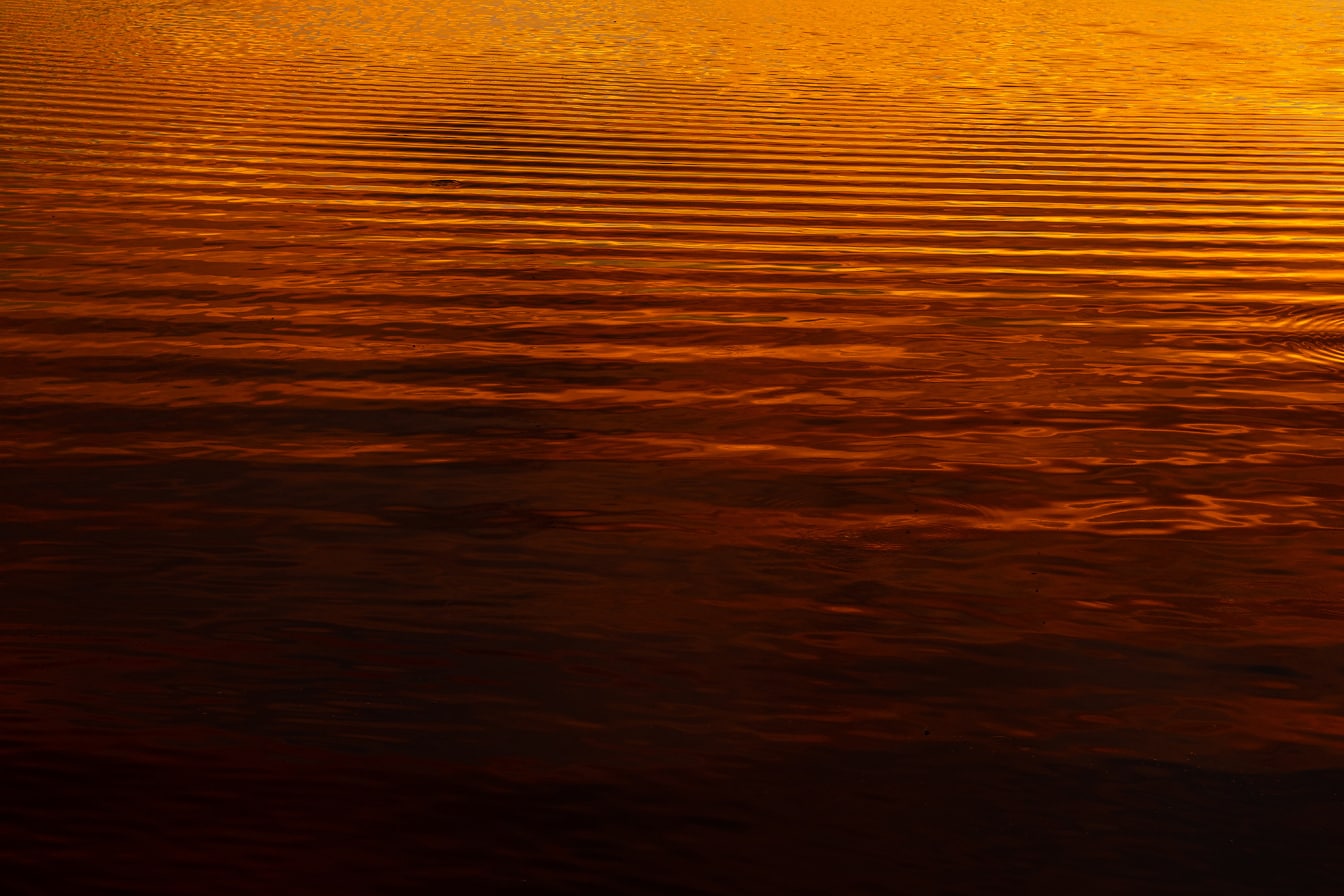 Tummanpunaisia ja oransseja keltaisia heijastuksia tyynen veden aalloilla auringonnousussa