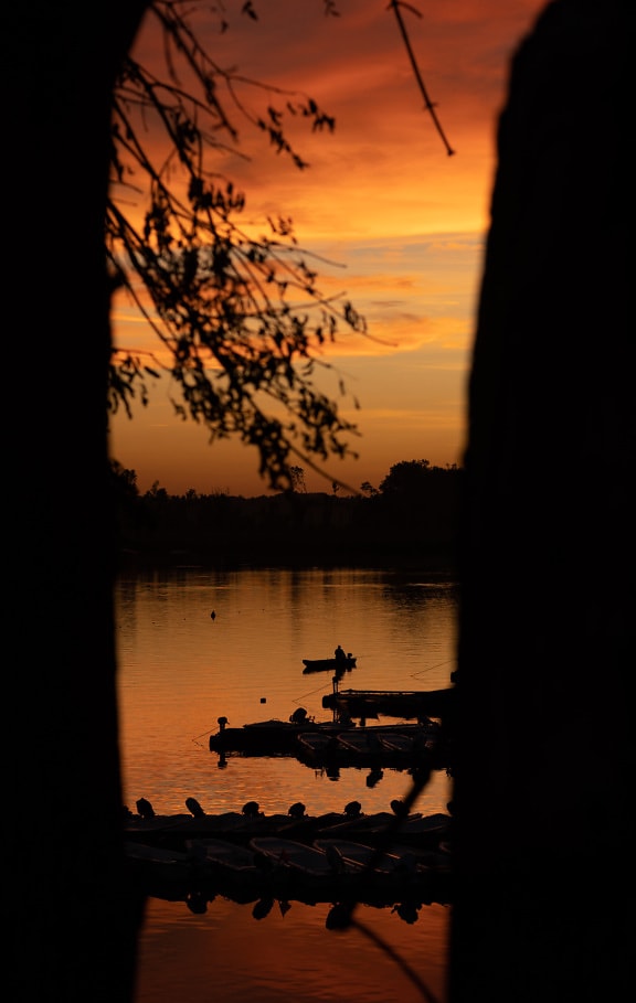 Silhouette of fishing boat in distance in orange yellow sunset on lakeside