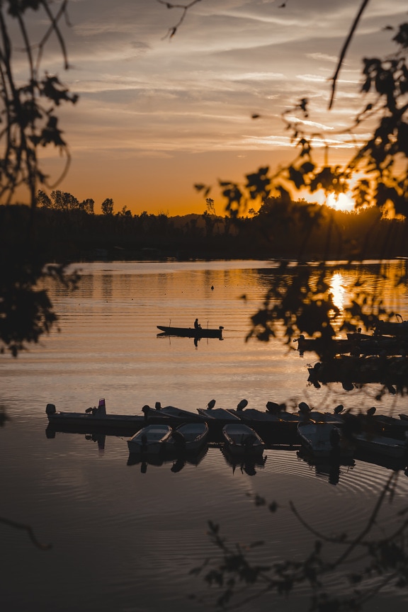 Pastelovo oranžovo žltý západ slnka so siluetou rybárskych lodí na brehu jazera
