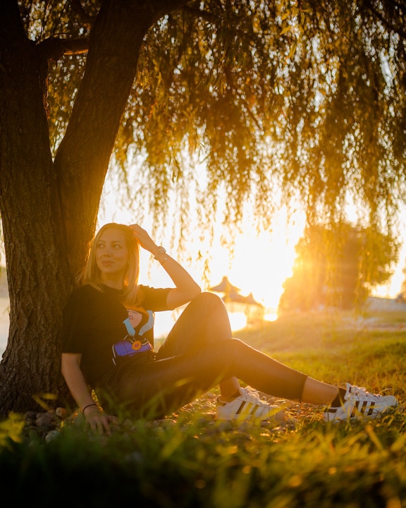 Smuk teenagerpige sidder under træet i solnedgang med solstråler i baggrunden