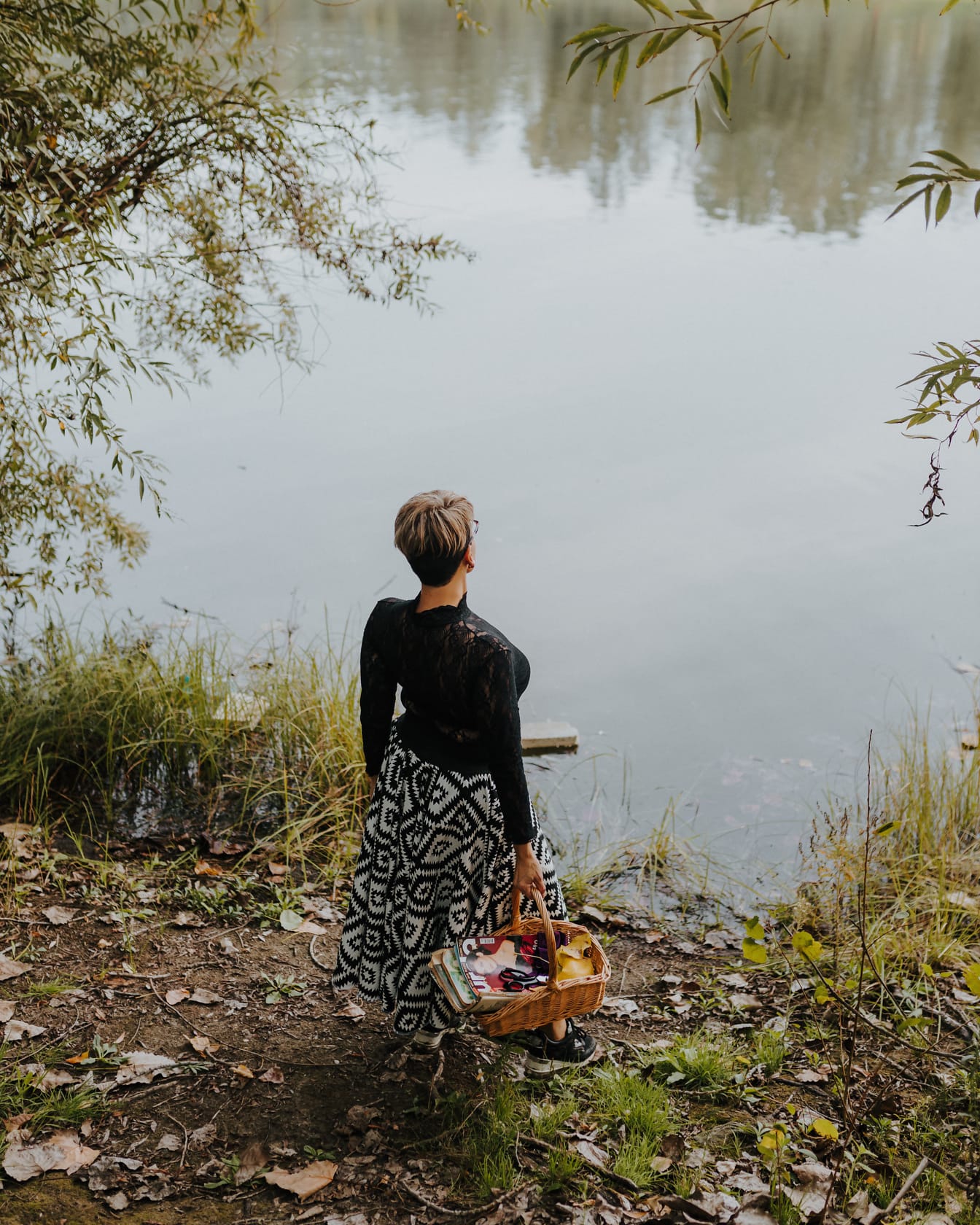 Молода жінка в чорно-білій сукні та плетеному кошику на березі річки
