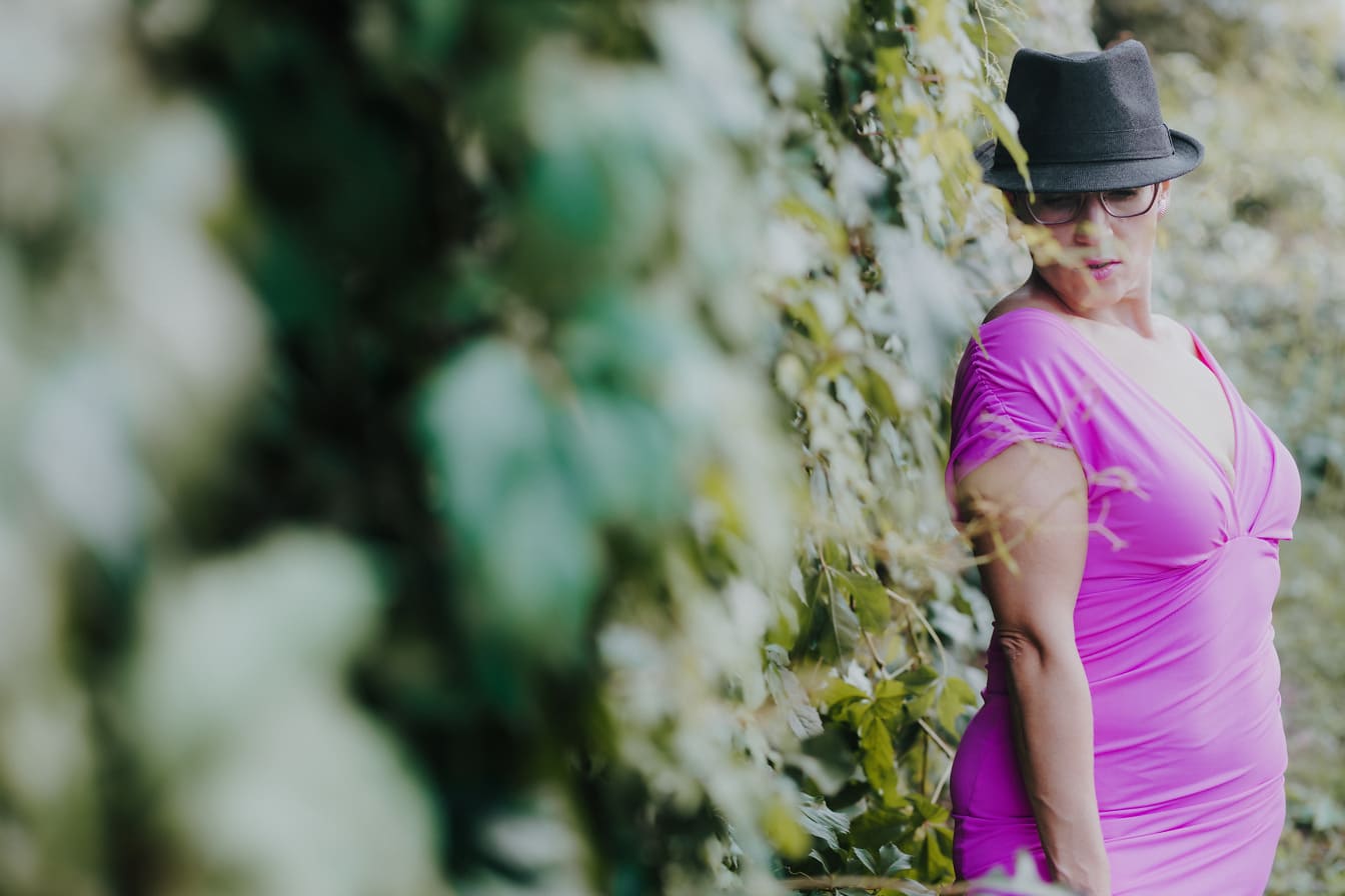 Kobieta nosi kapelusz i różowo-fioletową sukienkę przez bluszczowy żywopłot