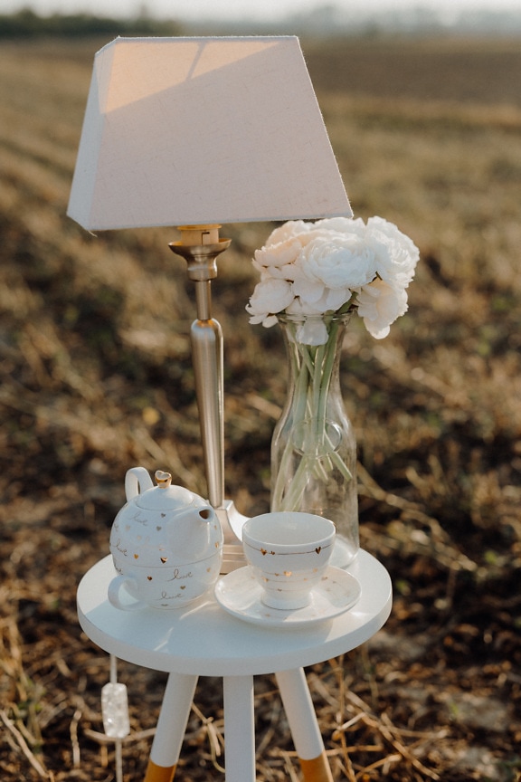 Lámpara de lujo con rosas blancas en jarrón y tetera sobre mesa en campo