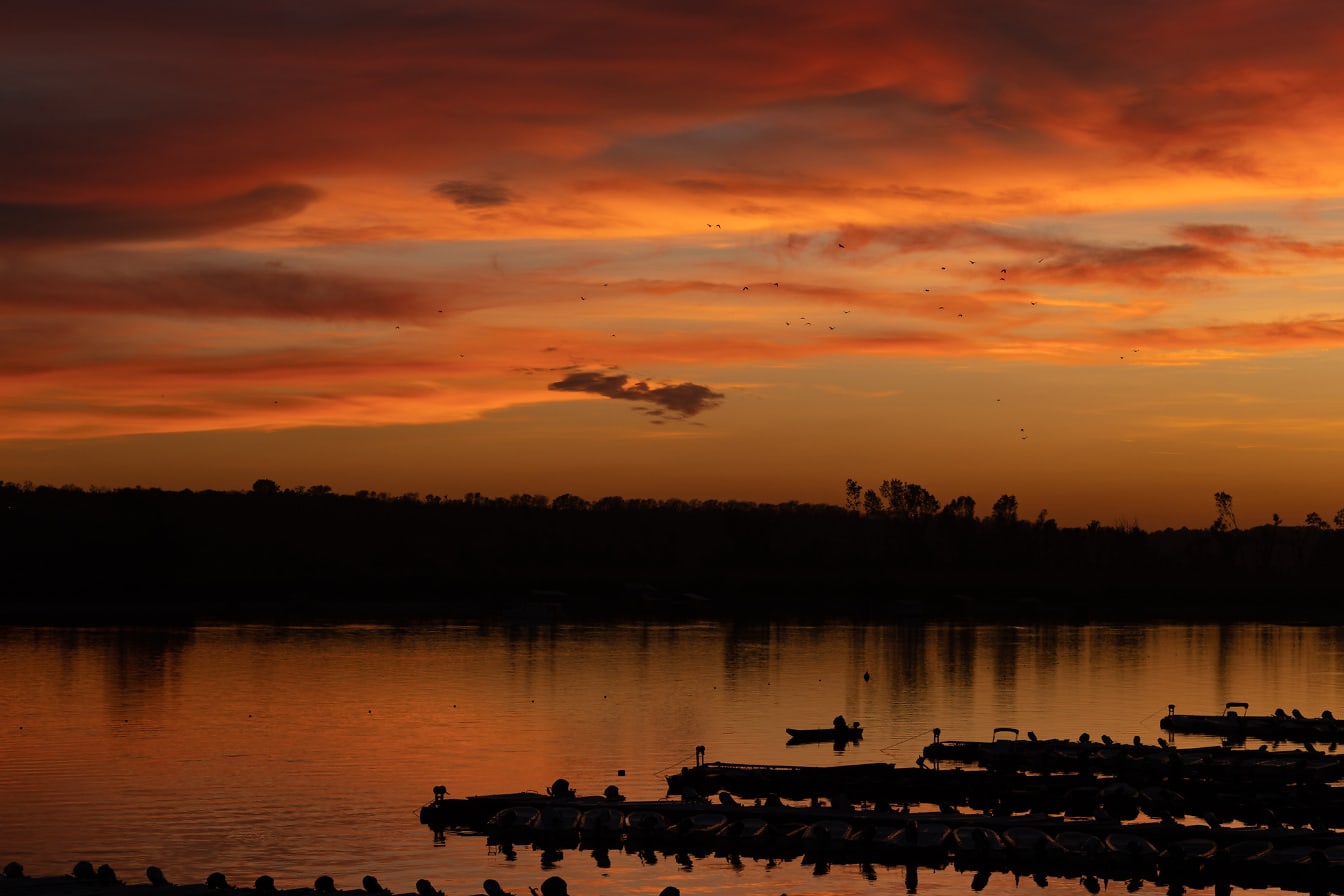 Ciemnoczerwone chmury zachodu słońca nad brzegiem jeziora z sylwetką łodzi rybackich