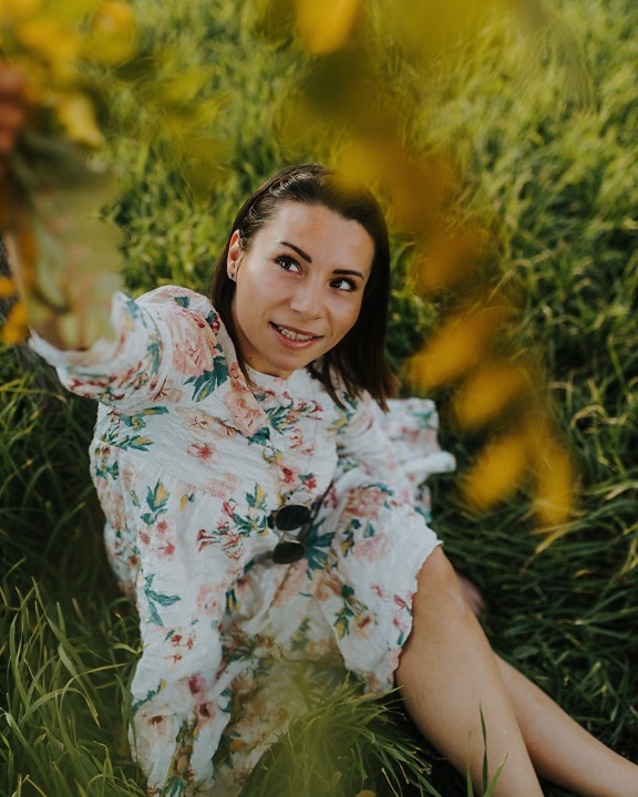 Nádherná brunetka sedí u trávy v okouzlujících květinových šatech