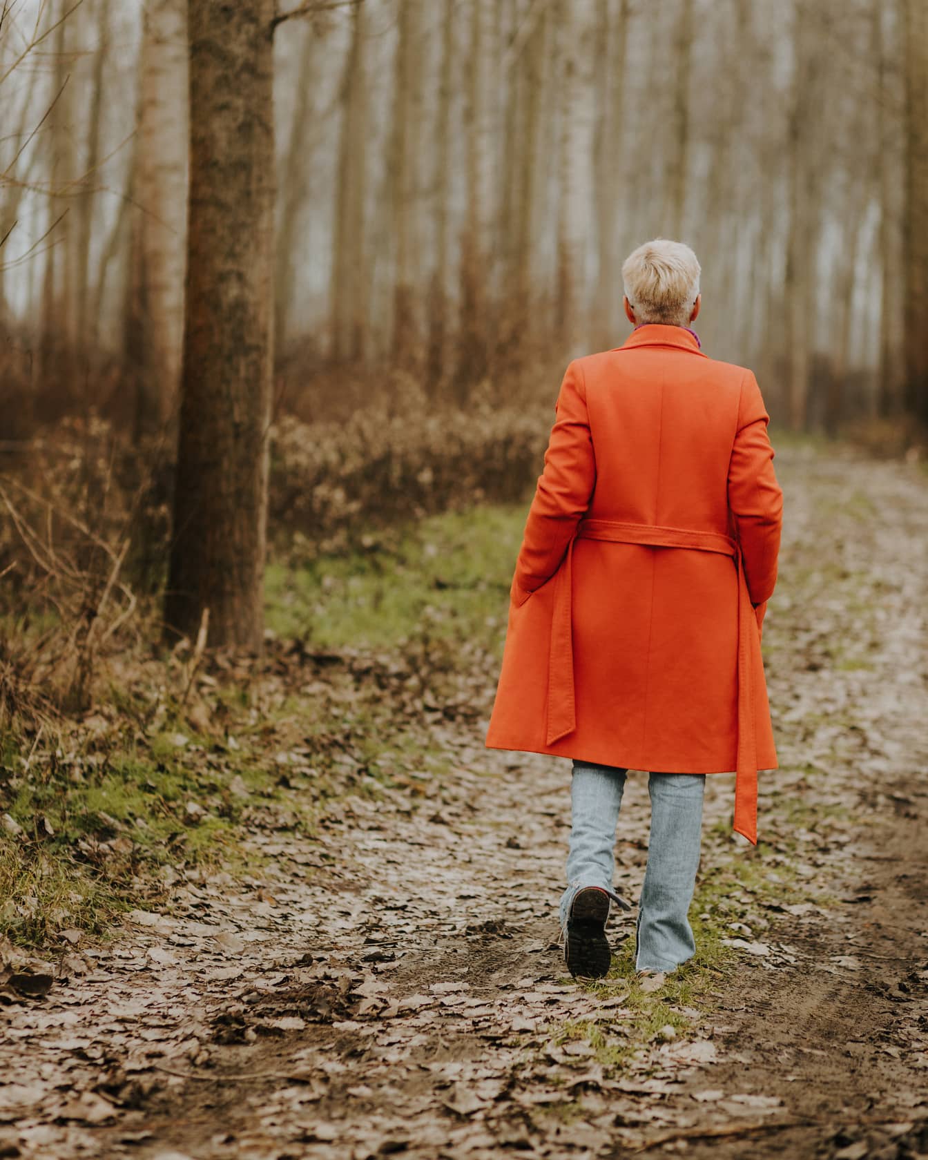 Kobieta w pomarańczowo-żółtym płaszczu spacerująca leśną ścieżką w jesienny dzień