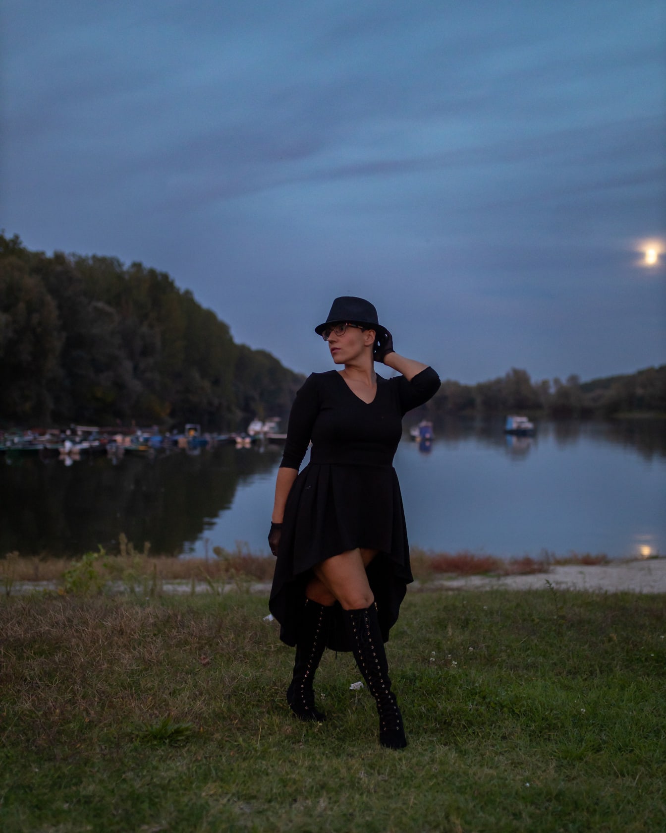 帅气的女人，戴着黑色帽子连衣裙和靴子，在湖边摆姿势