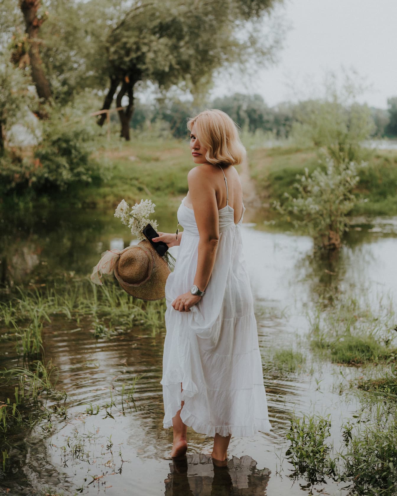 Prekrasna bosonoga plavuša u bijeloj haljini stoji u jezeru