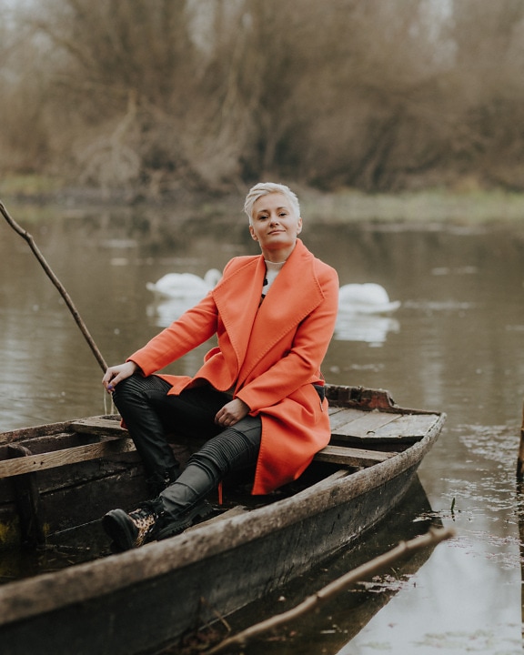 Fröhliche Frau in orangefarbenem Mantel und Lederhose sitzt im Boot