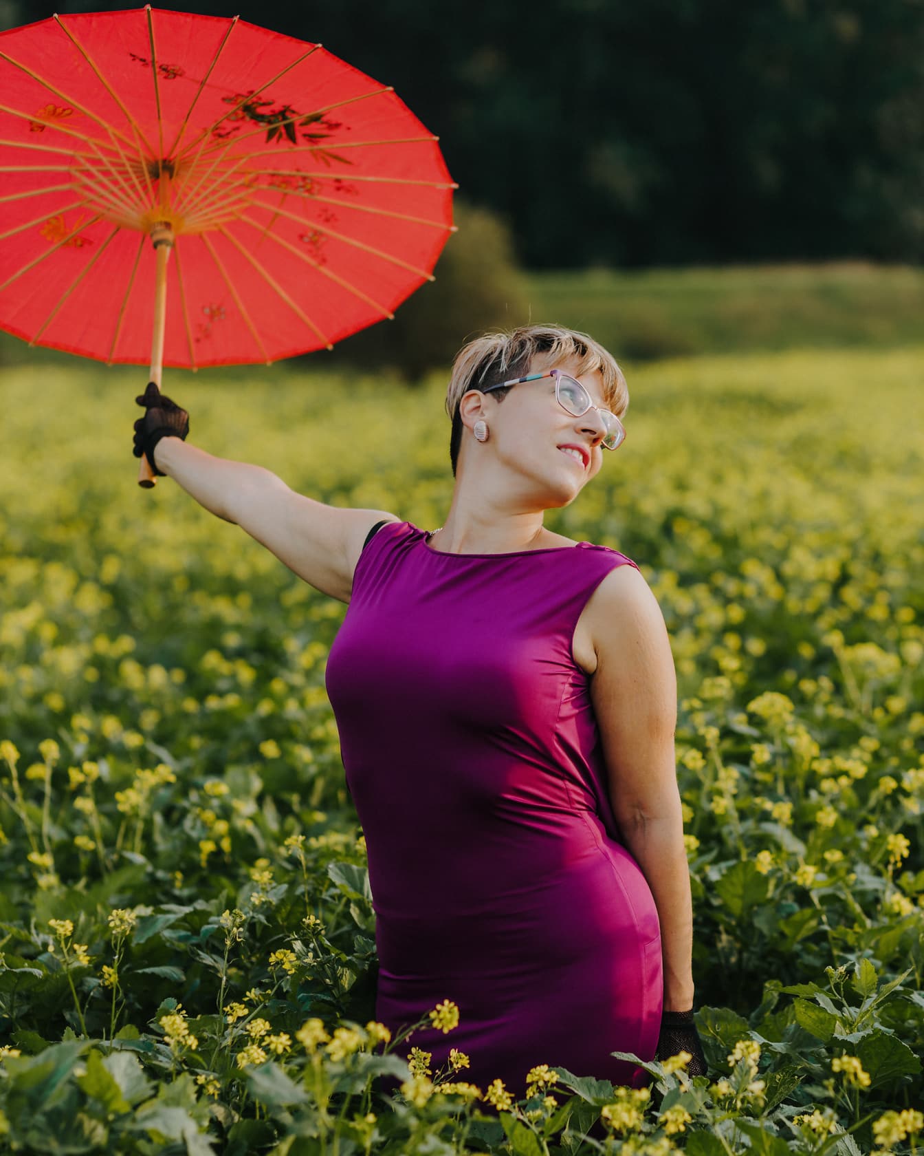 Blondă fericită cu umbrelă roșie în câmp de rapiță