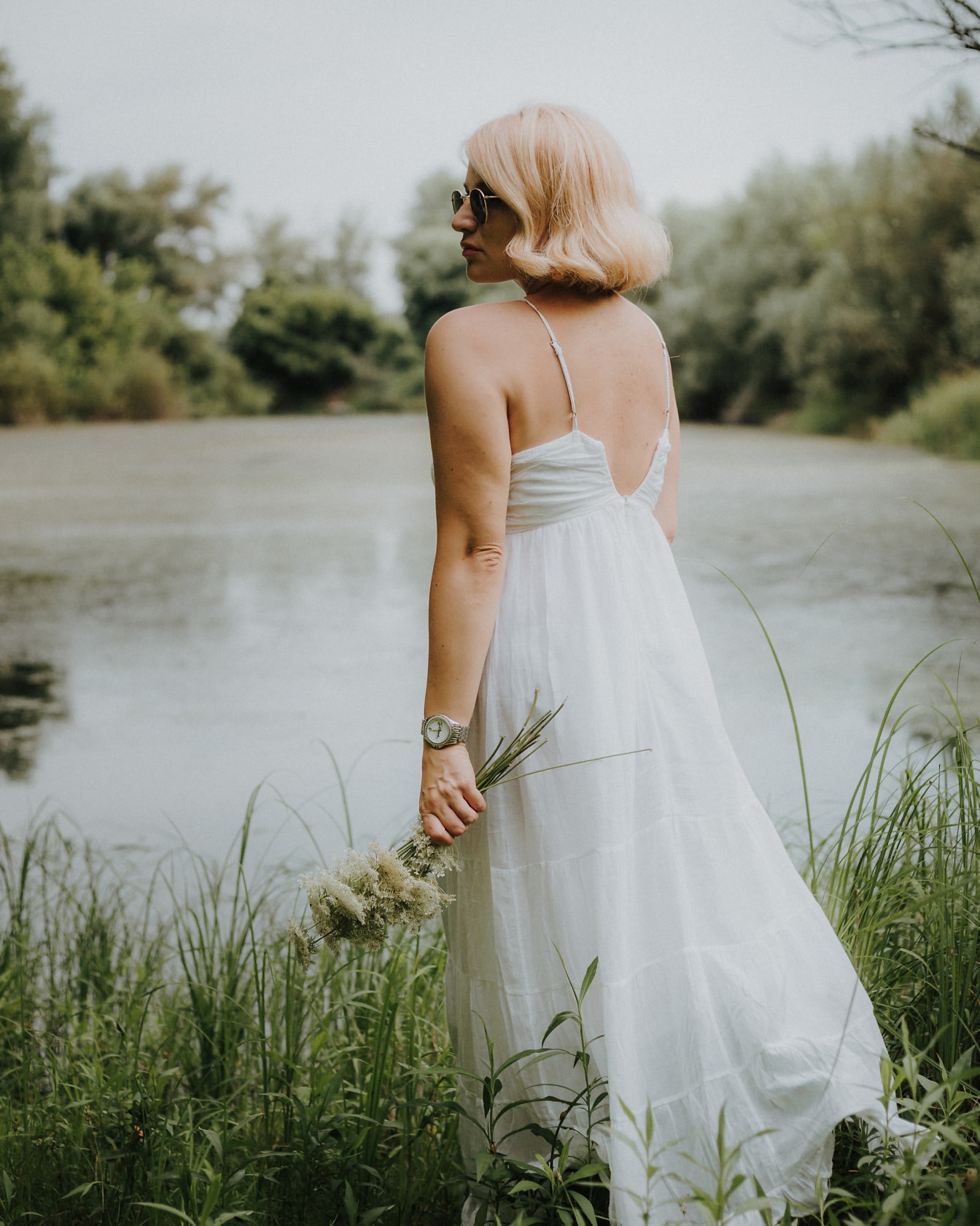 Блондинка з букетом квітів у білій сукні біля озера