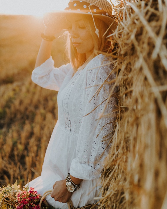 Người phụ nữ xinh đẹp với chiếc mũ tạo dáng trong đống cỏ khô với tia nắng mặt trời ở hậu cảnh