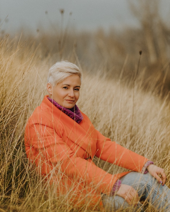 Hübsche blonde Frau posiert im trockenen Gras im orangefarbenen Mantel