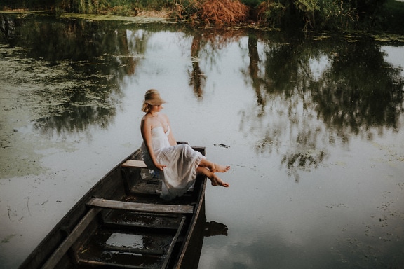 Barefoot woman in elegant white dress in rustic wooden boat on lake