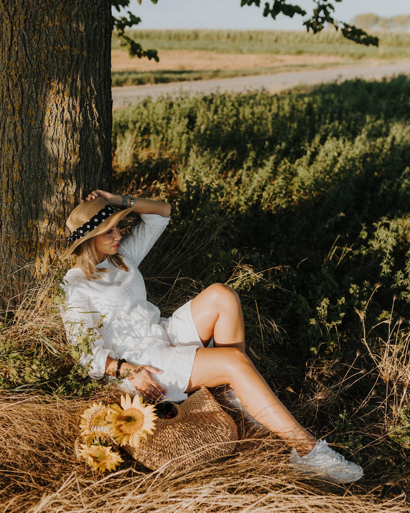 Ung kvinne som sitter i hvit kjole under treet på landsbygda