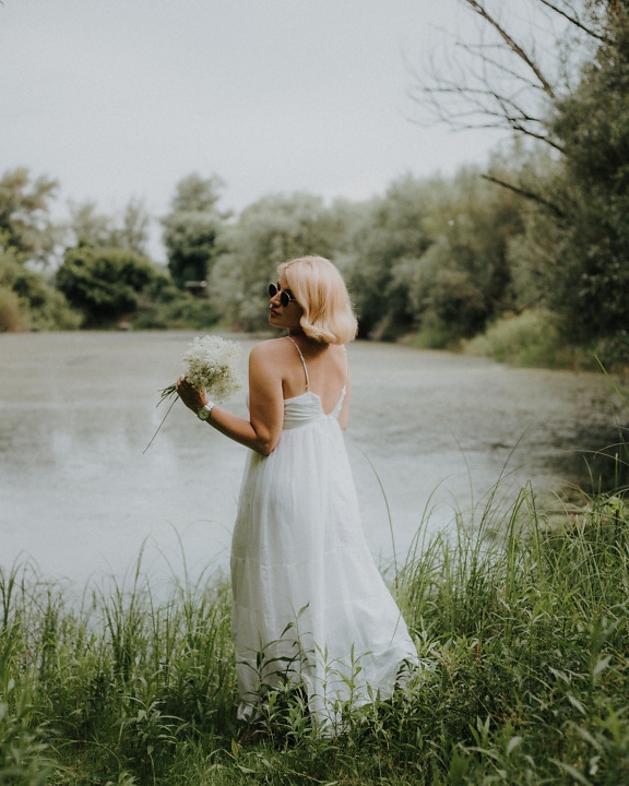 Beautiful blonde lady in elegant white dress holding flowers outdor