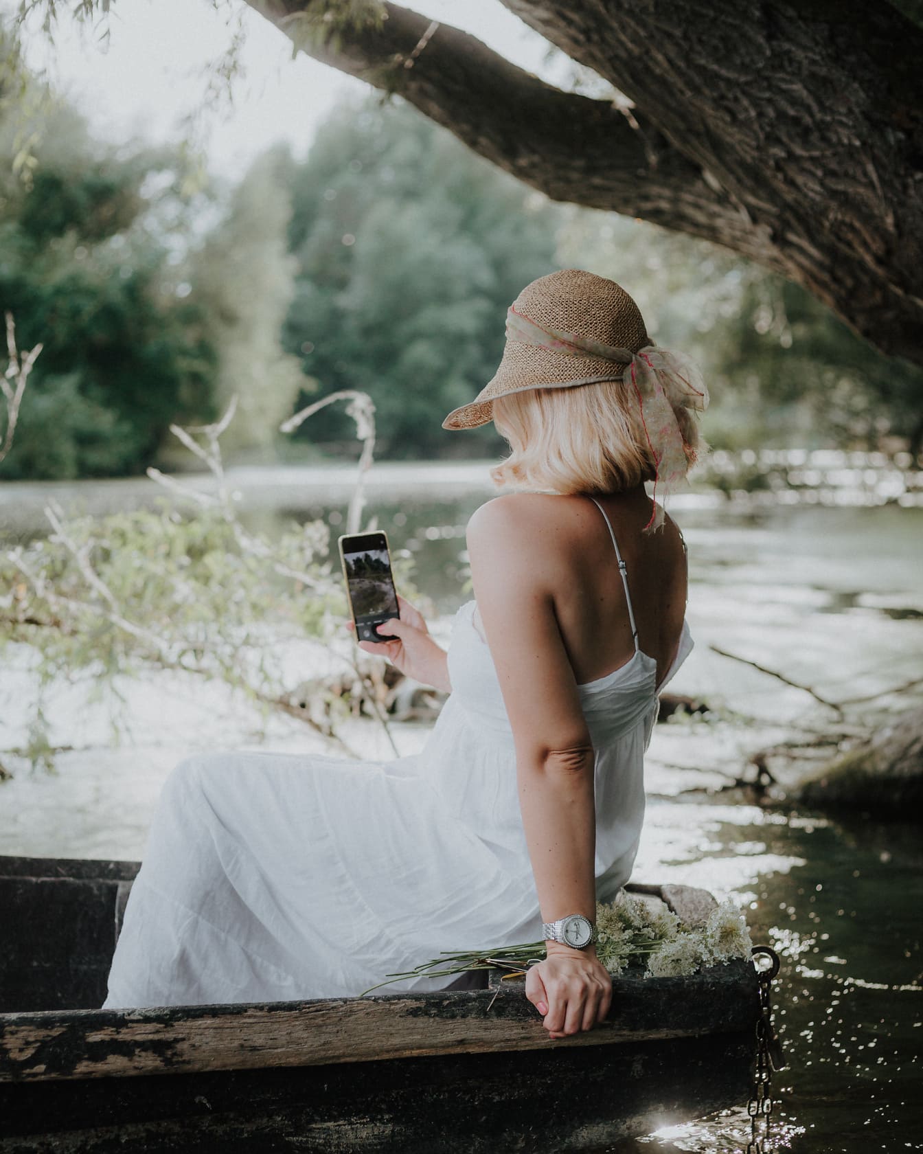 Блондинка в солом’яному капелюсі та білій сукні тримає мобільний телефон у сільській місцевості