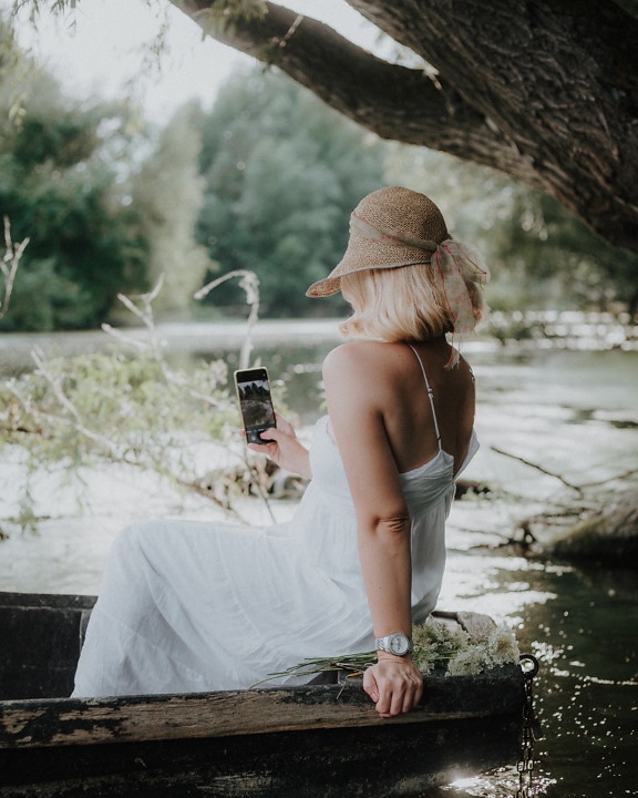 Blondýnka se slaměným kloboukem a bílými šaty drží mobil na venkově