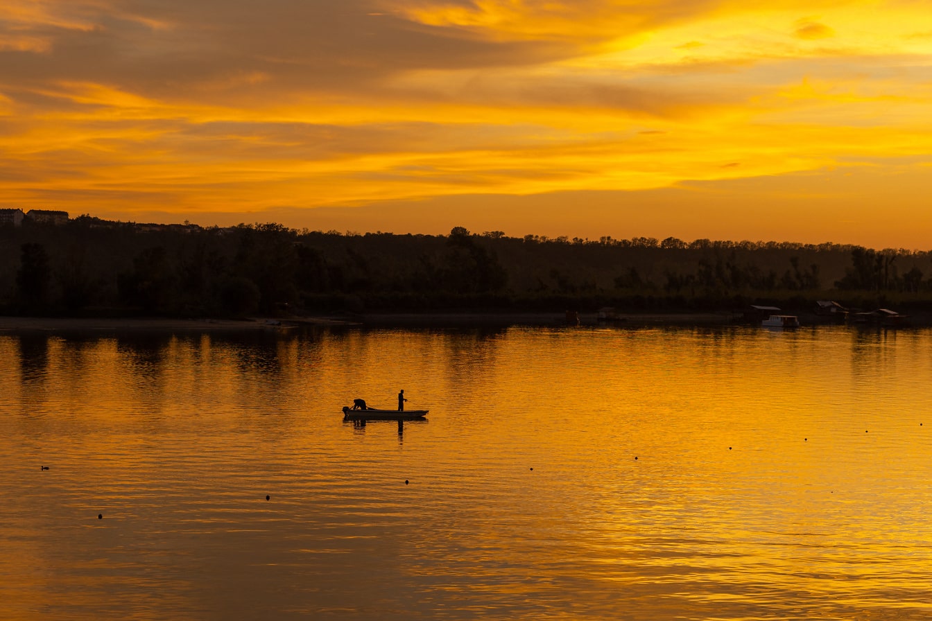 Nažloutlý východ slunce na břehu jezera se siluetou rybářské lodi