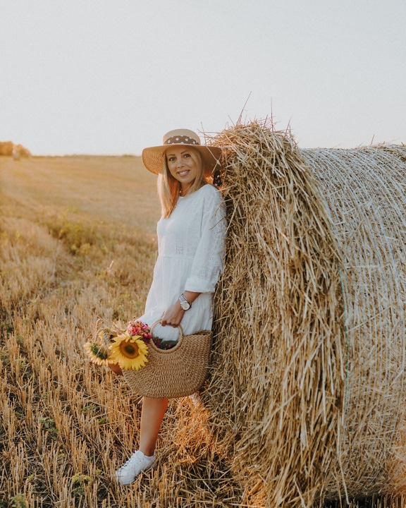 Veselá blondýnka se slaměným kloboukem v kupce sena v pšeničném poli za slunečného dne