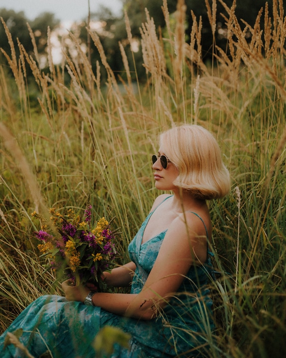 Piękna blondynka siedząca w trawie z bukietem kwiatów