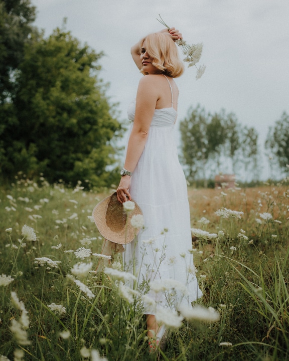 Добре изглеждаща блондинка, стояща в бяла рокля на поляна