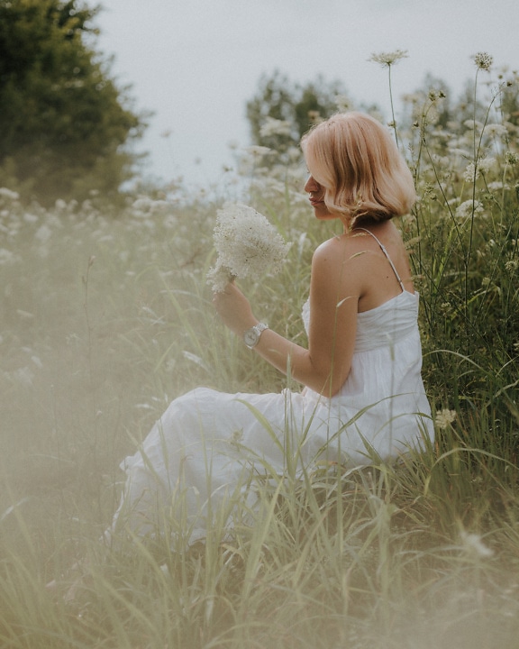 Samotna pani siedząca w wysokiej trawie łąkowej w białej sukience