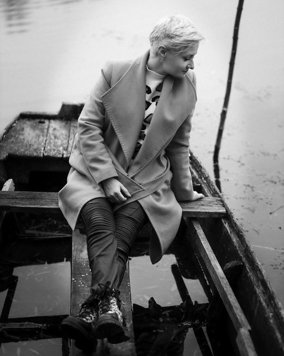 Monochrome photo of adorable blonde sitting in boat in coat