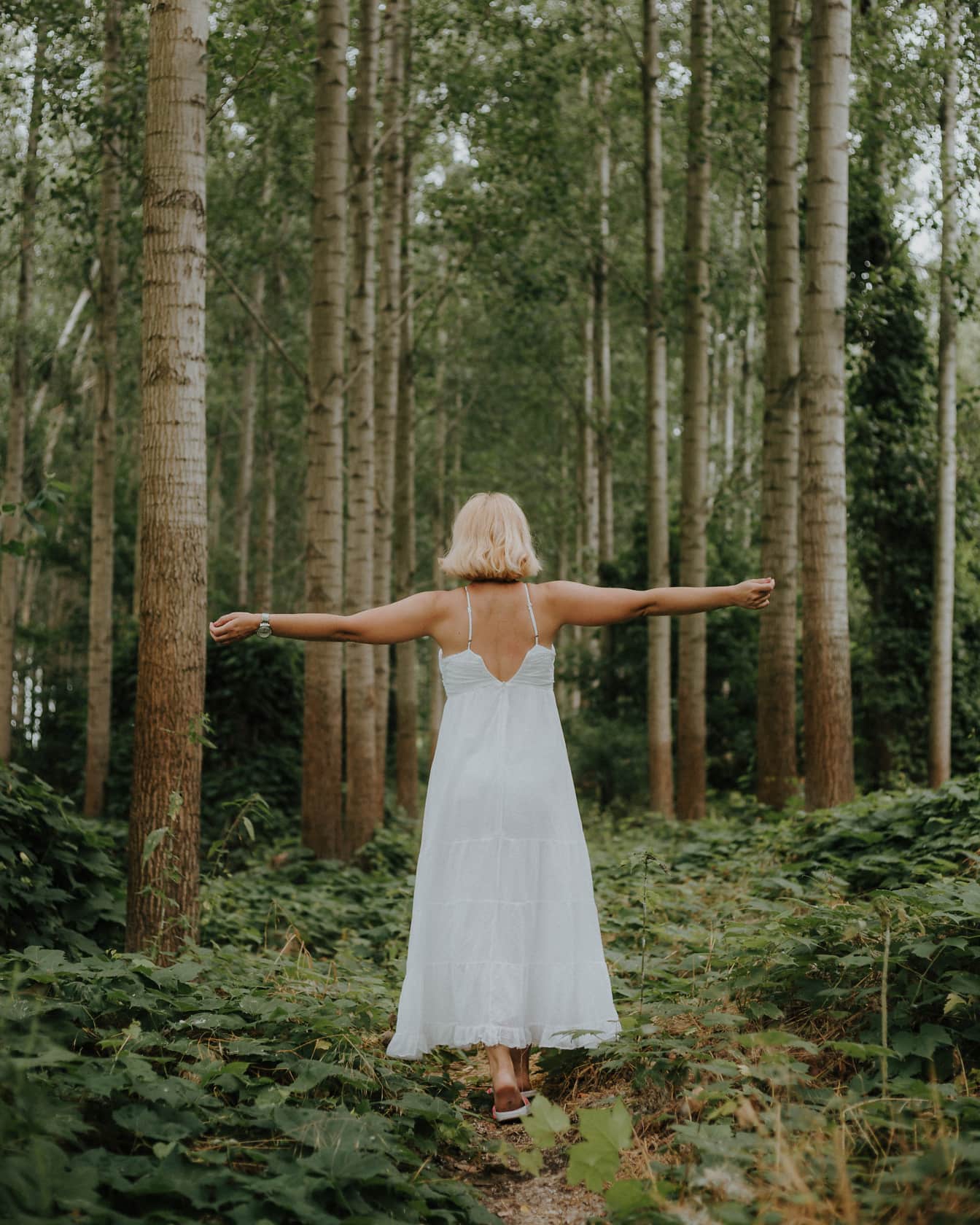 Blondynka stojąca w lesie ubrana w czystą białą sukienkę