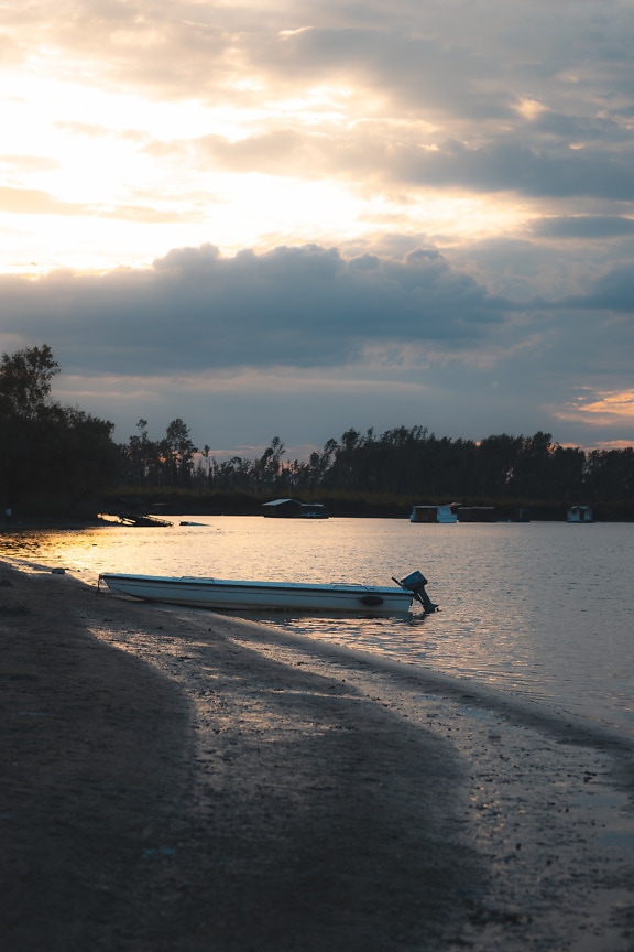 Bateau de pêche blanc sur la rive du lac de sable au crépuscule avec un ciel nuageux