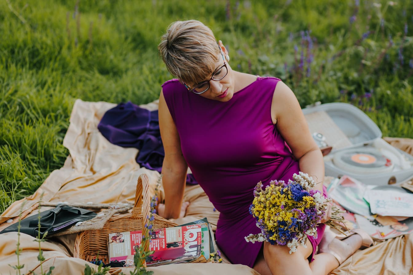 Dáma ve fialových šatech sedí na piknikové dece s kyticí květin