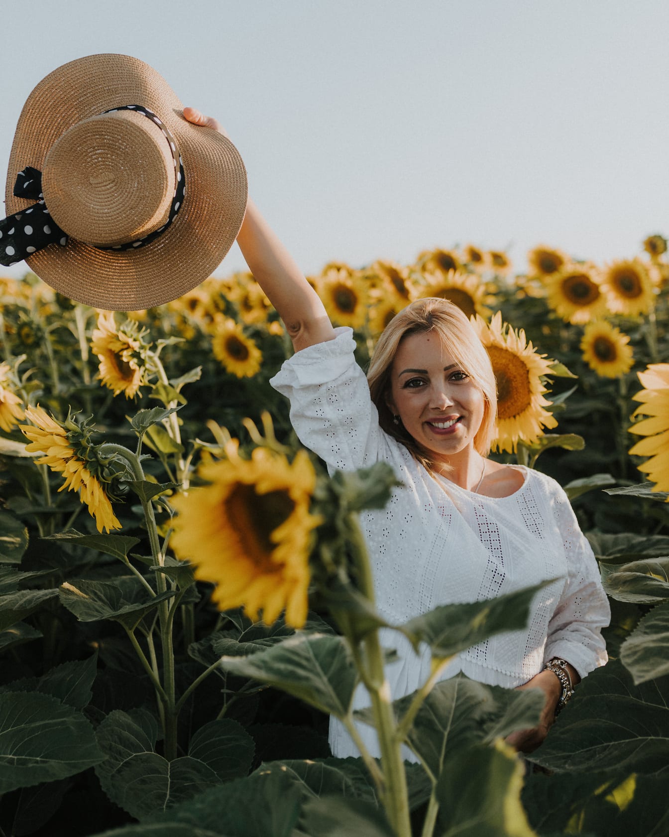 Nádherná blondínka usmievavá v slnečnicovom poli so slameným klobúkom v ruke