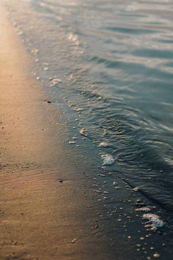Detail mokrého písku na pláži s odrazem slunečních paprsků na vlnách