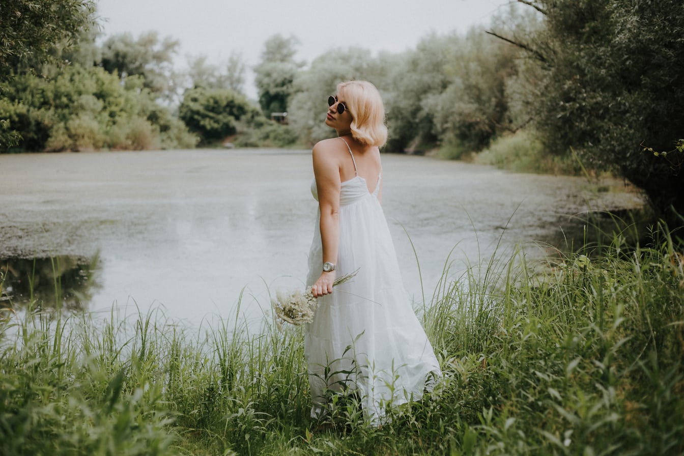 Blond panna młoda w okularach przeciwsłonecznych w sukni ślubnej na trawiastym brzegu jeziora