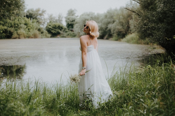 Blonďatá nevěsta se slunečními brýlemi ve svatebních šatech na travnatém břehu jezera