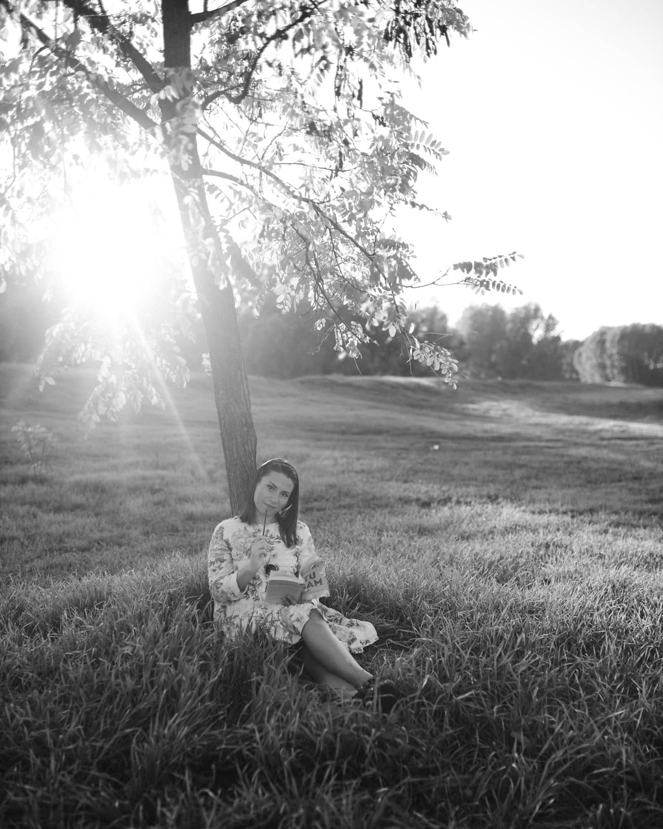 Monokromt portræt af ung kvinde, der sidder under træet og læser en bog