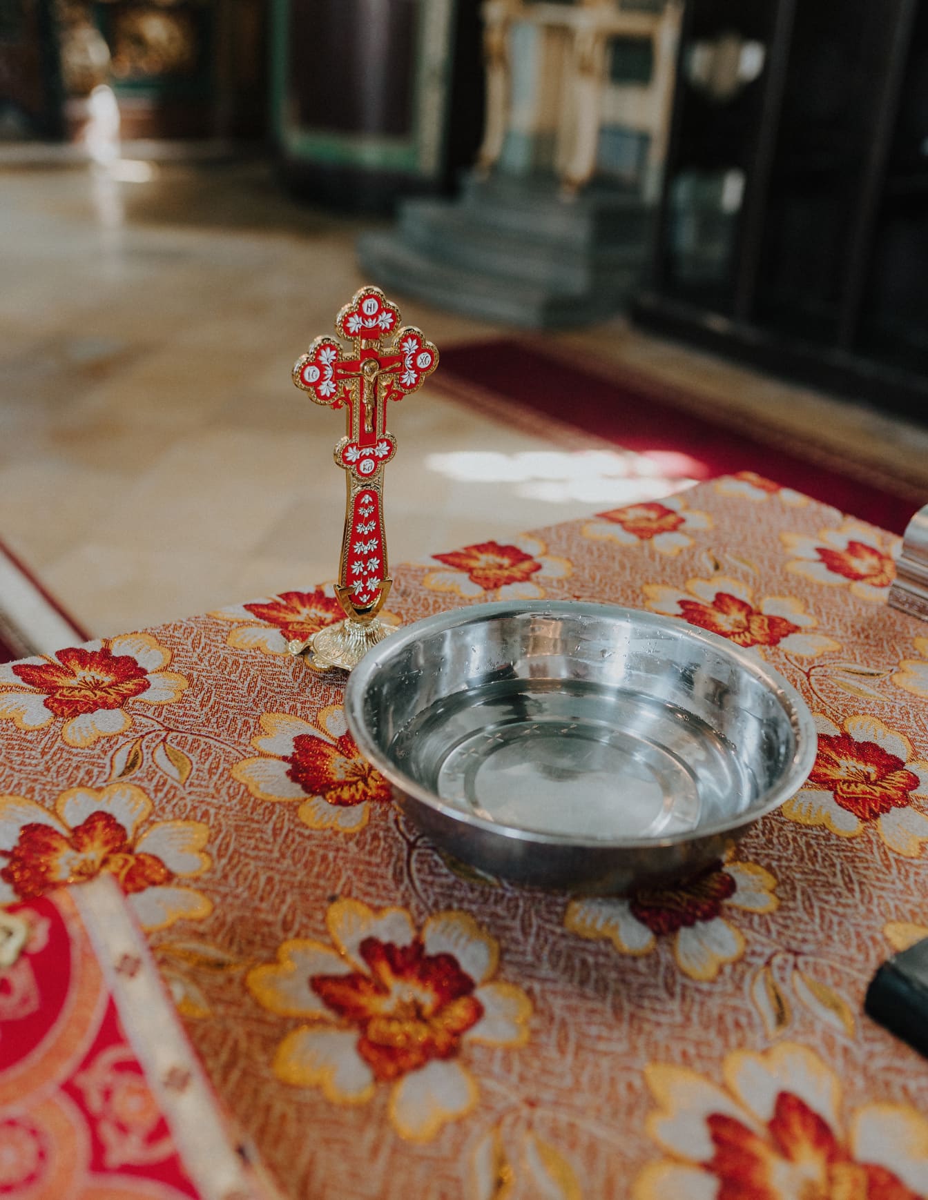 Cruce ortodoxă aurie cu ornament roșu închis și bol metalic pe masă