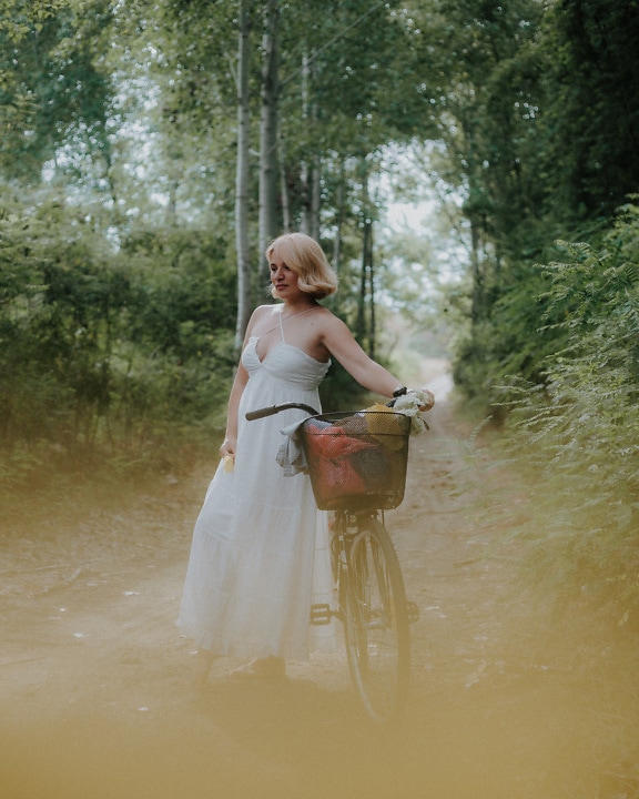 Hermosa rubia en el bosque con la bicicleta en el camino del bosque